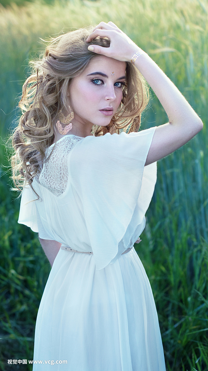 诱人的金发碧眼的女孩,蓝色的眼睛站立在白色的衣服在高大的草地